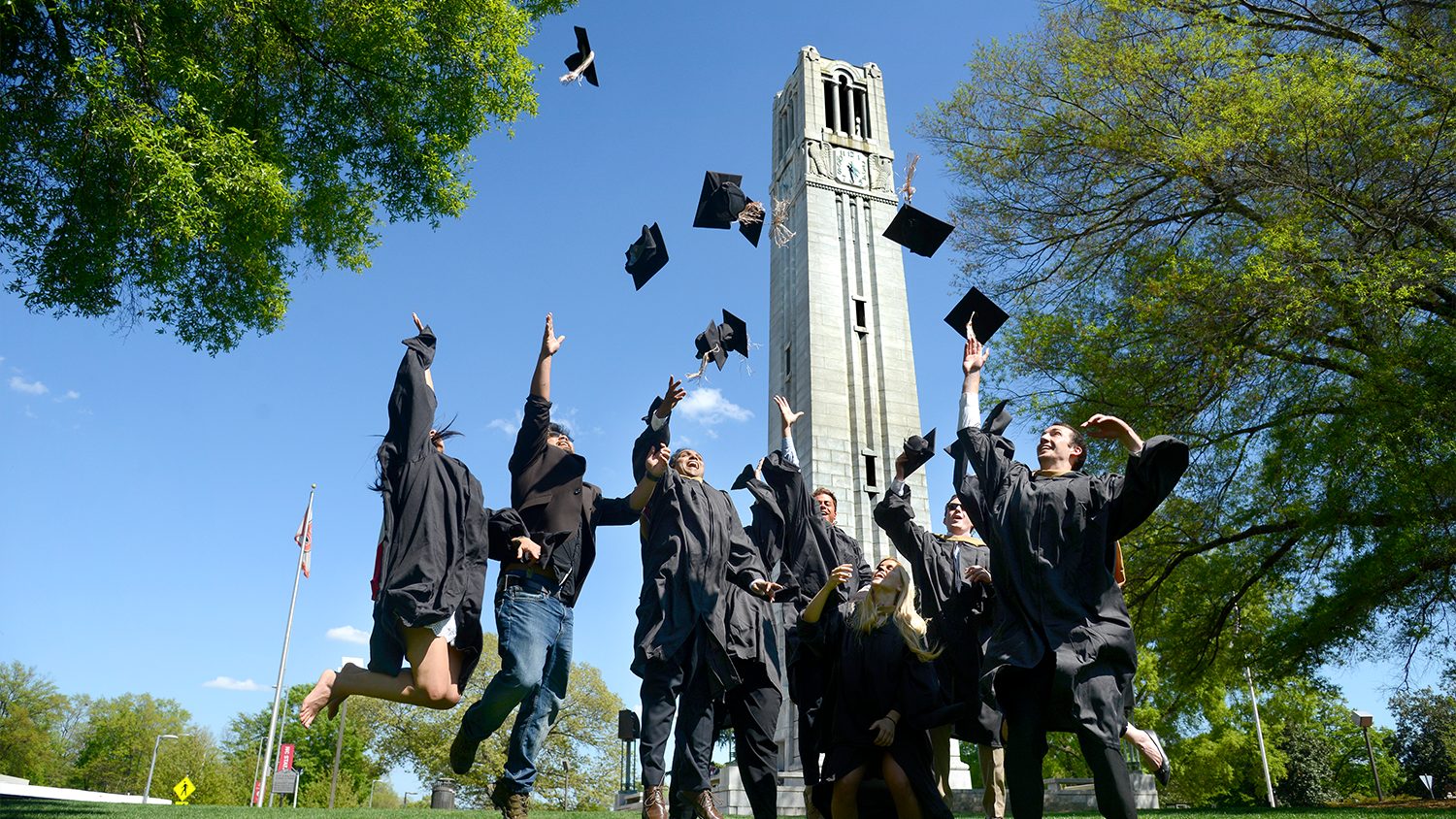 A group photo of Master students who are jumping for air in their black cap and gown in front of the NC State belltower.