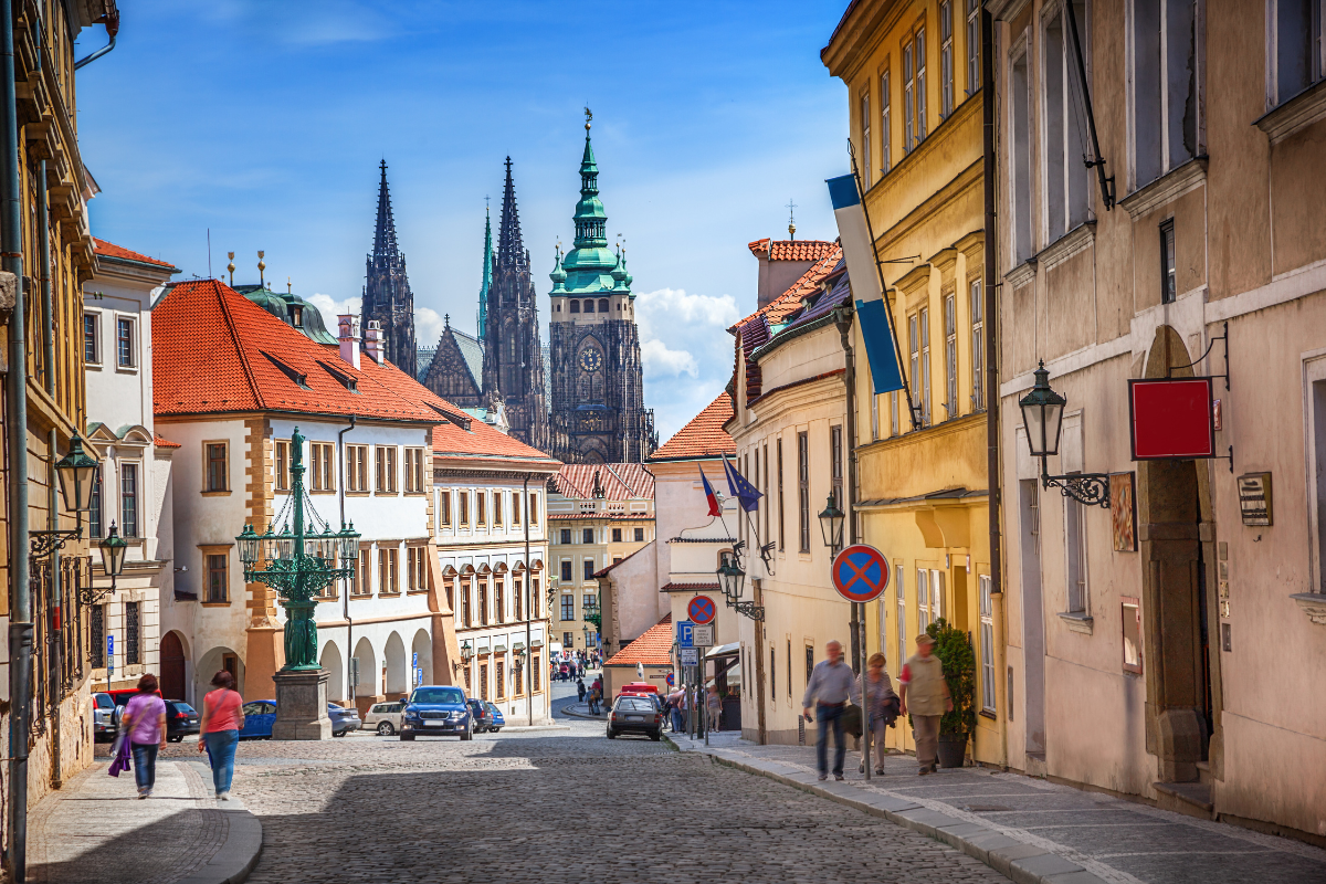An image of Prague with a cathedral in the background against a blue sky with people walking down the street. 