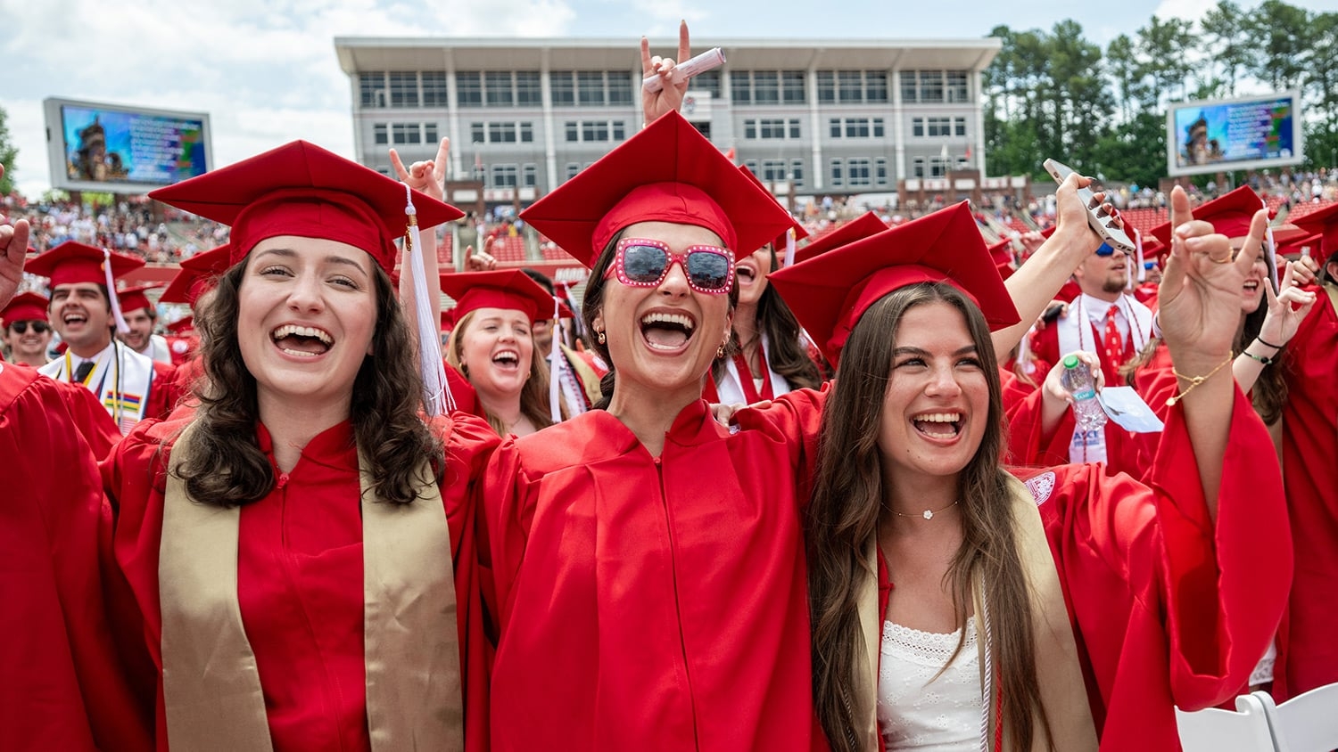 Three graduates in red caps and gowns cheer during commencement.