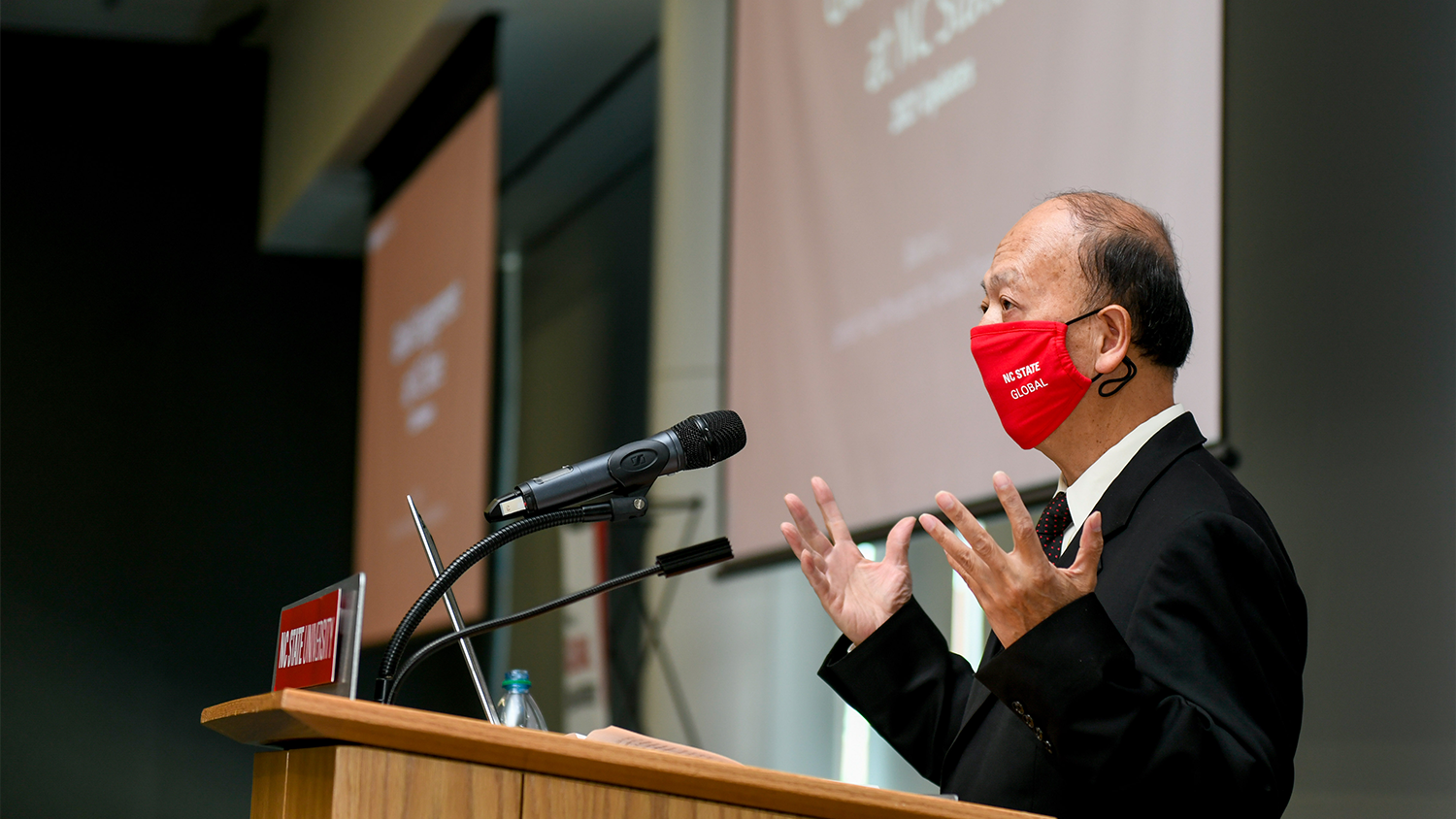 A man with a mask on giving a speech at a podium in front of a big screen.