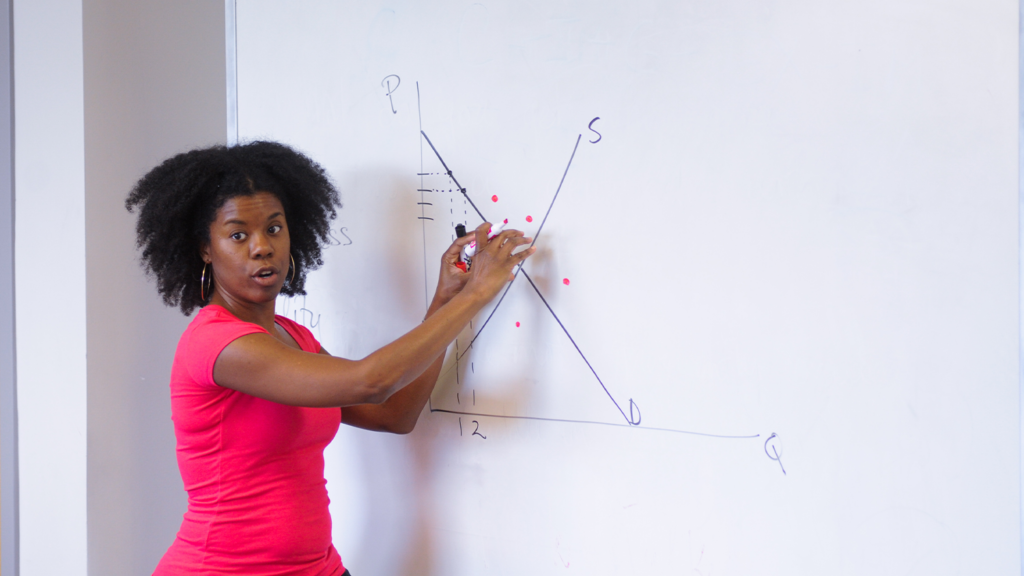 A young black female professor is standing at a whiteboard explaining supply and demand.