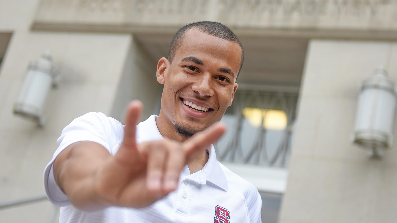 A young black man in a white NC State polo shirt making a wolf sign with his hands.