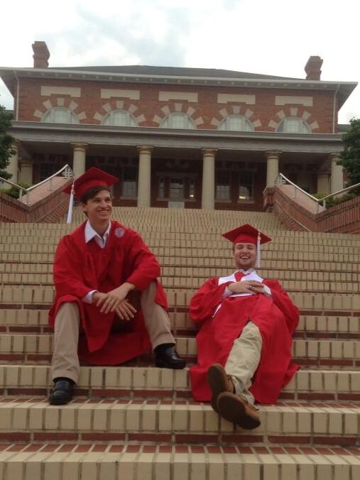Jimmy Pagett with a friend taking graduation pictures in their red undergraduate gown on the steps of the 911 building in the Court of Carolinas.