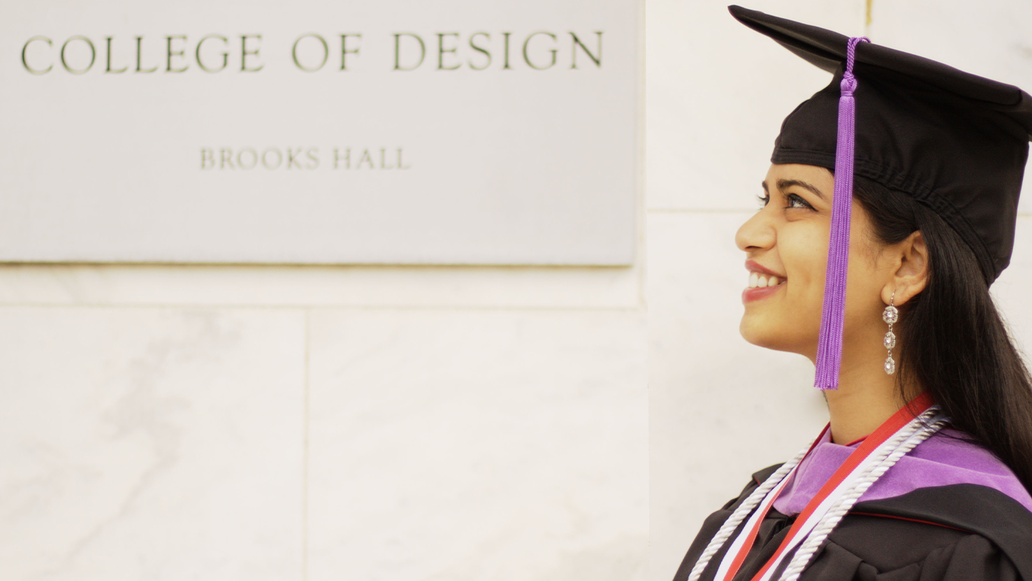 A graduate in their gown standing next to the College of Design.